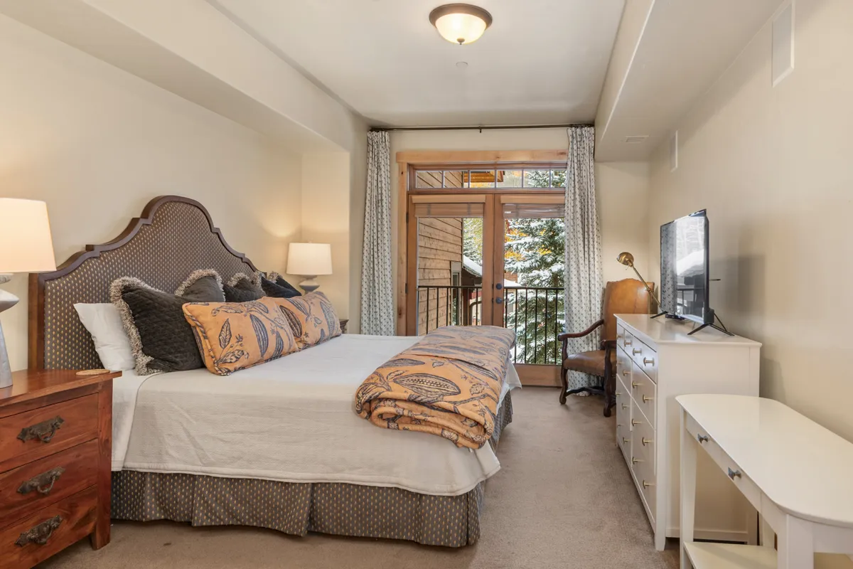 A spacious bedroom with a king-size bed adorned with decorative pillows, a large window offering access to a balcony, and a television atop a dresser. Soft morning light fills the room, creating a tranquil atmosphere ideal for relaxation.