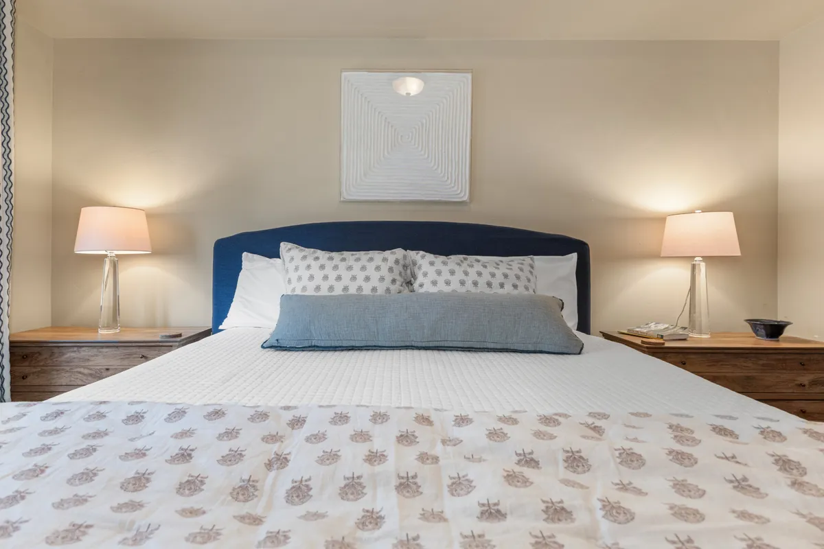 A cozy bedroom scene with a neatly made bed accentuated by decorative pillows, framed wall art, and softly glowing bedside lamps on nightstands. The room radiates serenity and is thoughtfully arranged for comfort and relaxation.