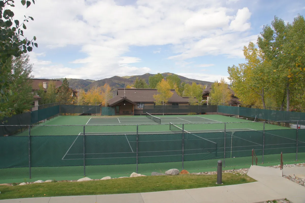 An exterior shot featuring tennis courts surrounded by lush trees with a striking mountain backdrop. The scene captures energetic tennis matches in progress and hints at the active, social lifestyle available to residents. The sunset light adds vibrancy to the scene, emphasizing outdoor recreation.