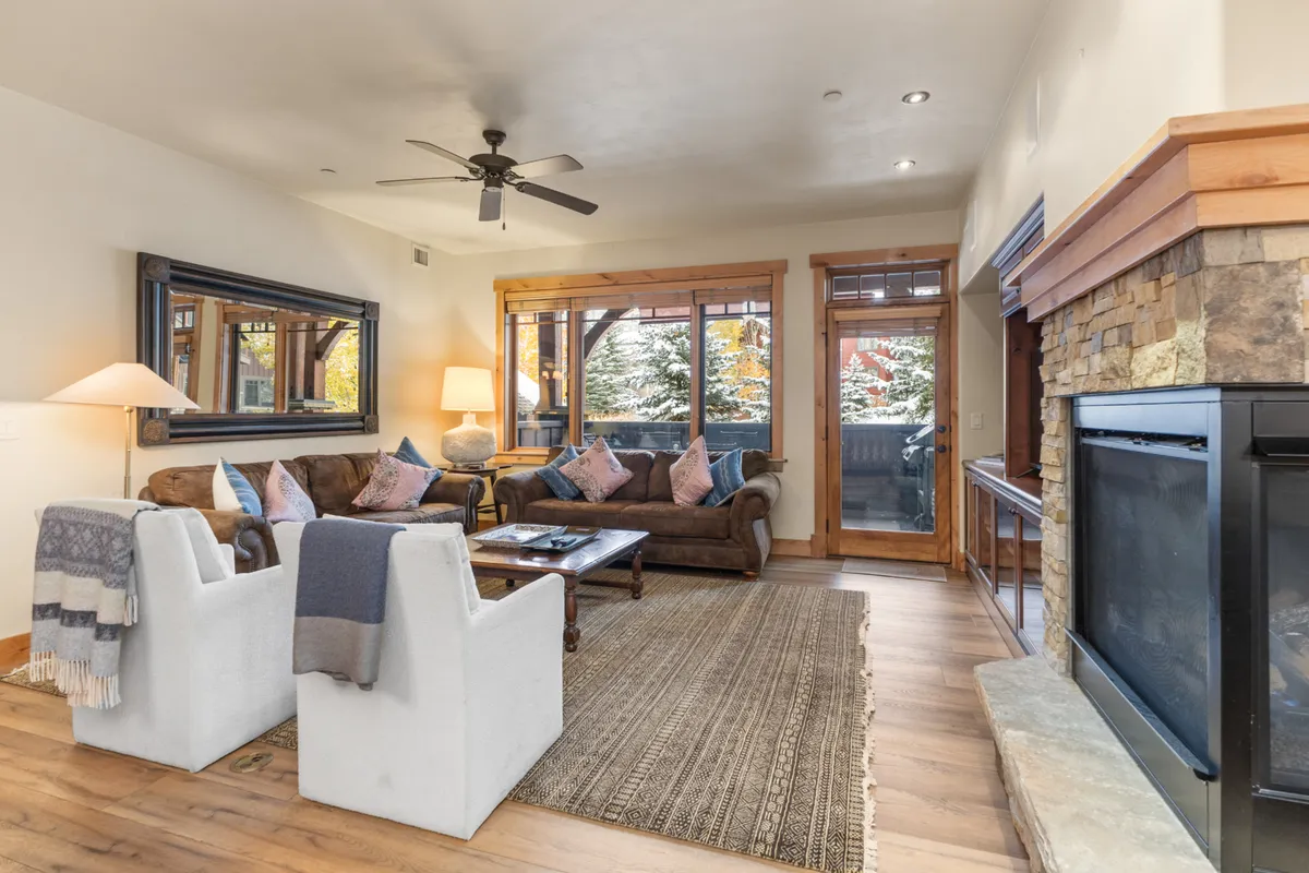 A spacious, welcoming living room centers around a glowing fireplace, with a plush sofa, coffee table, and large windows that let in abundant natural light. The arrangement fosters conversation and comfort, with warm decor touches and a sense of openness.