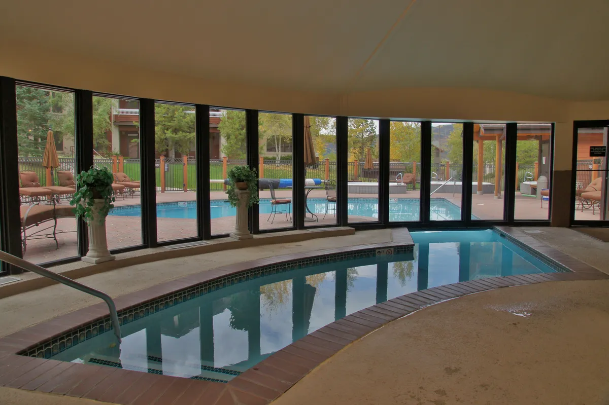 The interior features an elegant indoor hot tub with large windows that overlook a swimming pool, flooded with natural light and decorated with lush plants. Comfortable lounge seating suggests relaxation and wellness, all within a serene, spa-like atmosphere.