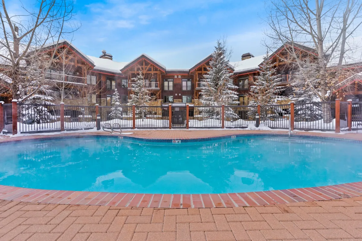 This exterior image features a serene swimming pool surrounded by trees and the lodge’s facade, with hints of social spaces where guests can relax or mingle. The ambiance is cozy and communal, suggesting opportunities for group relaxation after a day of exploring or skiing.