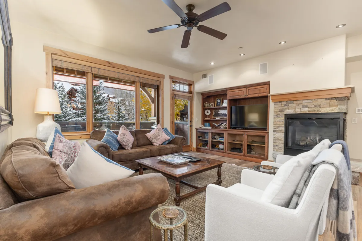 The image showcases a warm, inviting living room with a plush sofa and coffee table centered around a glowing fireplace and TV. Large windows flood the space with natural light and offer glimpses of mountain scenery, hinting at the ski-in/ski-out convenience. The arrangement encourages relaxation, movie nights, and cozy gatherings after a day on the slopes.