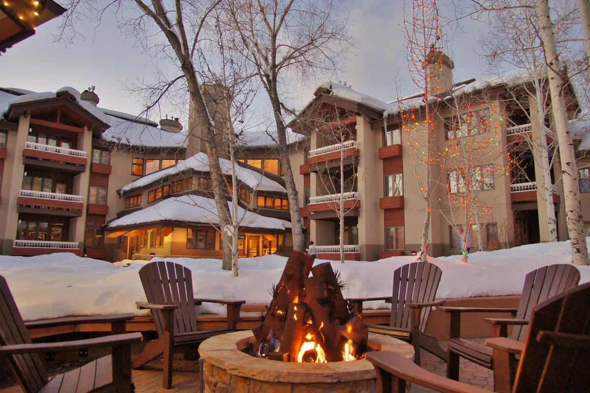 The image focuses on a welcoming fire pit area with surrounding chairs, charming building architecture, and snow-dusted ground. Decorative lights on nearby trees add a festive, communal mood, perfect for family gatherings after mountain adventures.